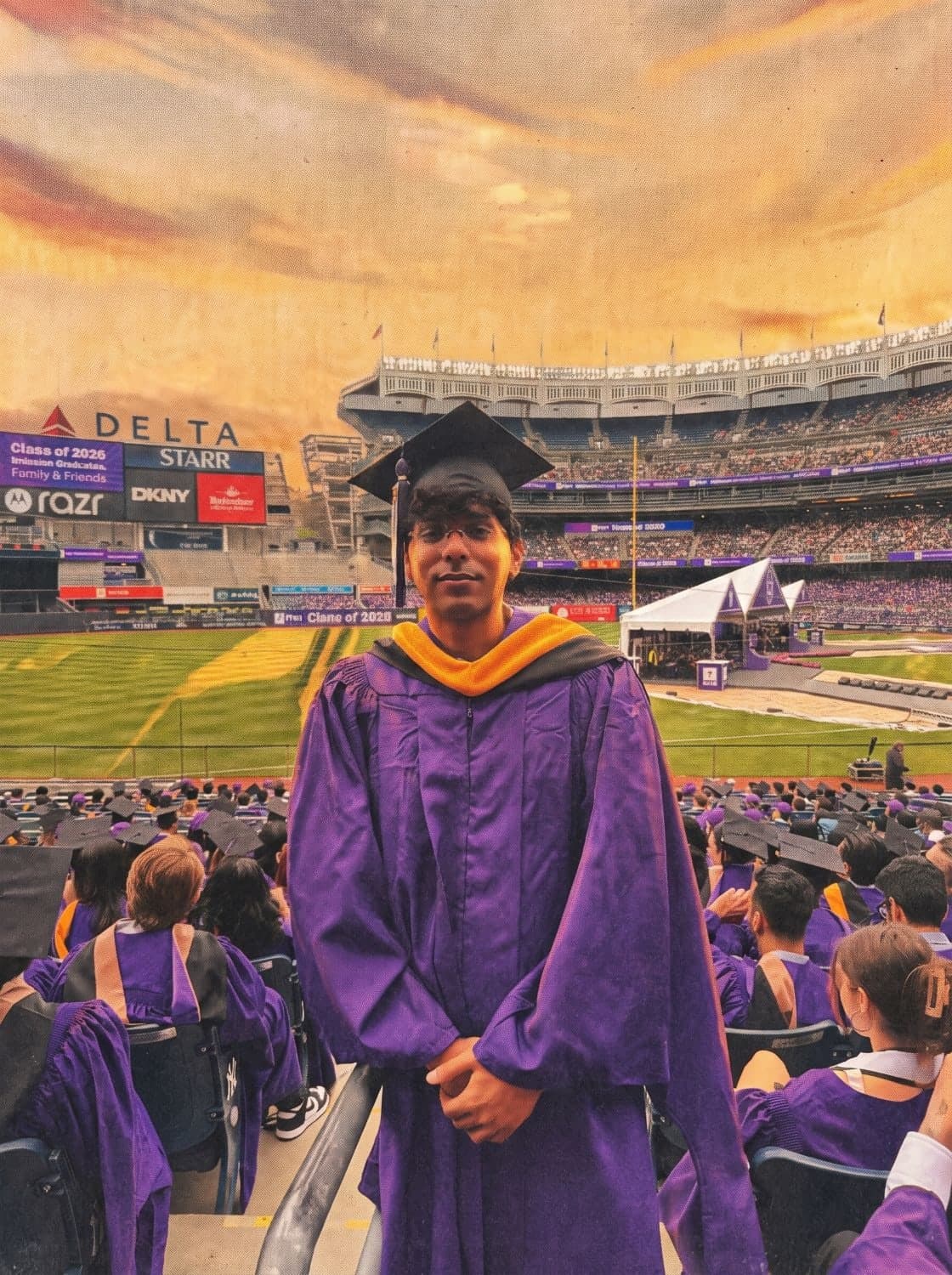 Yash Nirwan in an NYU violet gown and mortarboard at the Class of 2026 commencement in Yankee Stadium, the field and a stage tent behind him